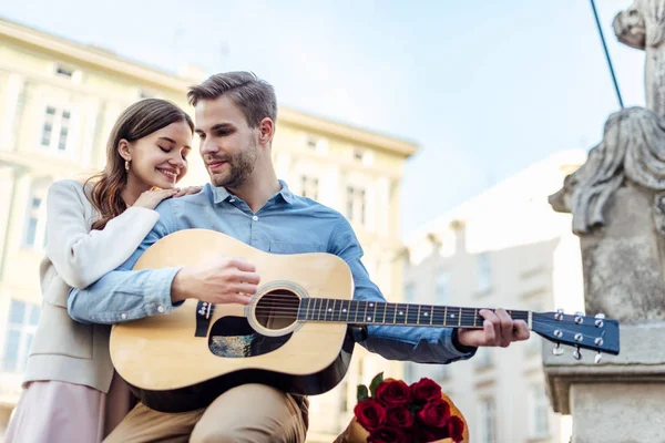 Glückliches Mädchen, das sich auf die Schulter seines Freundes lehnt und auf der Straße Akustikgitarre spielt — Stockfoto