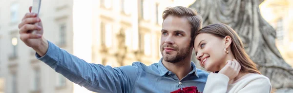 Panoramic shot of handsome man taking selfie on smartphone with happy girlfriend — Stock Photo
