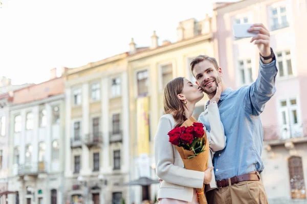 Mujer joven sosteniendo ramo de rosas y besando novio tomando selfie en el teléfono inteligente - foto de stock