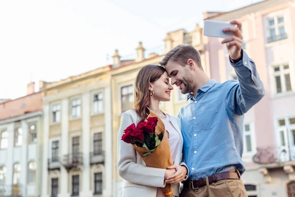 Heureuse jeune femme tenant bouquet de roses tout en souriant petit ami prendre selfie sur smartphone — Photo de stock
