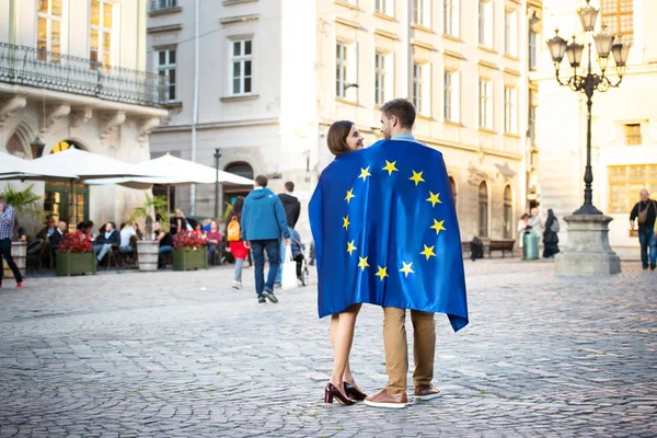 Couple de jeunes touristes, enveloppé dans le drapeau de l'union européenne, debout sur la place de la ville — Photo de stock