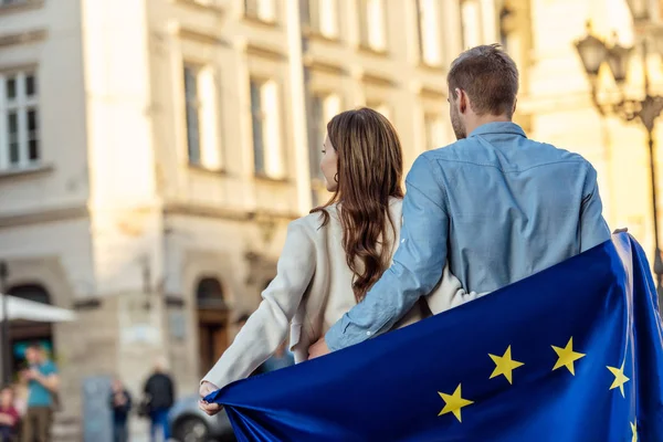 Vista trasera de la pareja joven, envuelta en la bandera de la unión europea, de pie en la calle - foto de stock