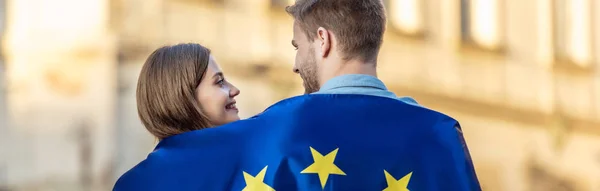Plano panorámico de joven pareja de turistas, envueltos en la bandera de la unión europea, mirándose unos a otros en la calle - foto de stock