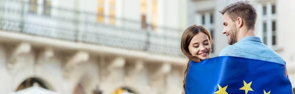 Panoramic shot of young, smiling tourists wrapping in flag of european union on street — Stock Photo