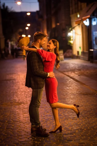 Feliz, elegante chica sosteniendo ramo de rosas mientras abraza novio en la calle de la noche - foto de stock
