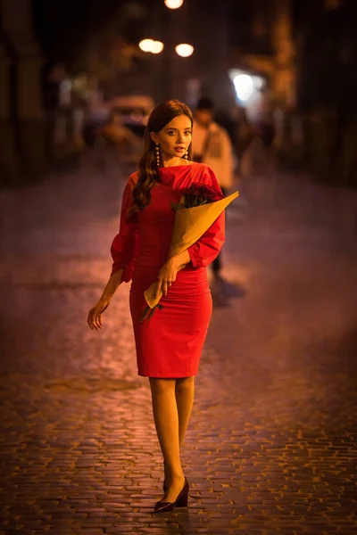 Attraktive junge Frau im eleganten roten Kleid mit einem Strauß Rosen, während sie nachts auf der Straße spaziert — Stockfoto