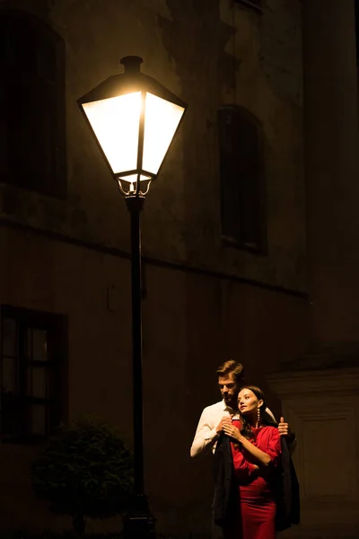 Joven hombre cubriendo congelado novia con chaqueta mientras de pie bajo farola en la noche - foto de stock