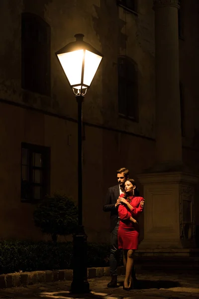 Joven abrazando elegante, novia congelada de pie debajo de la lámpara de la calle por la noche - foto de stock