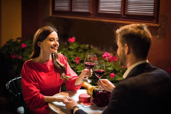 Chica feliz tintineo vasos de vino tinto con novio haciendo propuesta de matrimonio en el restaurante - foto de stock