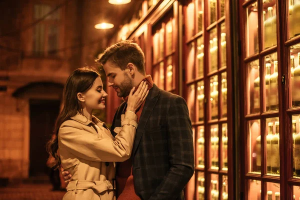 Happy girl touching handsome boyfriend whlie standing near shop window at night — Stock Photo