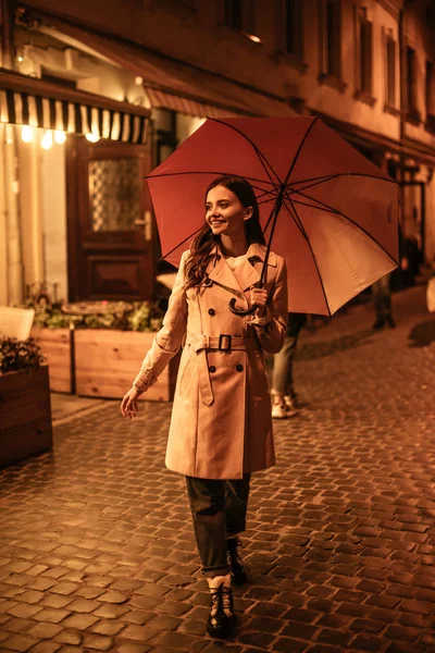 Fröhliche junge Frau spaziert mit Regenschirm auf der Abendstraße — Stockfoto