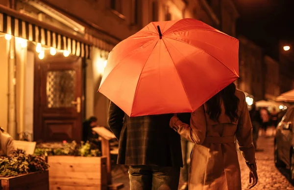 Rückansicht eines Pärchens im herbstlichen Outfit, das unter einem Regenschirm die Abendstraße entlangläuft — Stockfoto