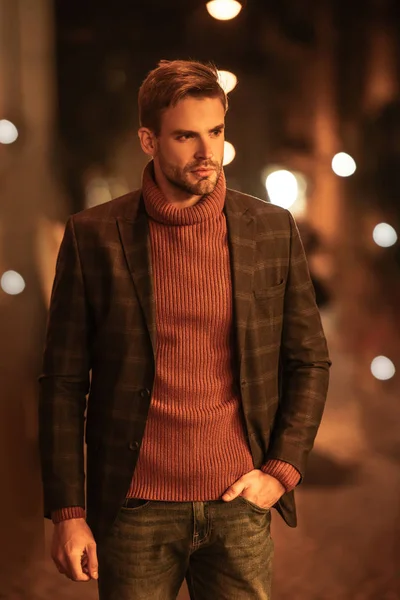 Hombre guapo y seguro en traje de otoño mirando hacia otro lado y tomándose de la mano en el bolsillo mientras está de pie en la calle de la noche - foto de stock
