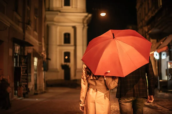 Vista trasera de la pareja en traje de otoño caminando bajo paraguas a lo largo de la calle de la noche - foto de stock