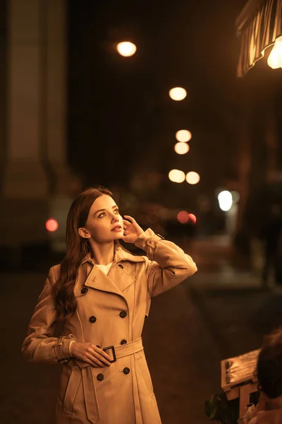 Chère jeune femme en trench coat regardant loin et touchant les cheveux tout en se tenant debout sur la rue du soir — Photo de stock