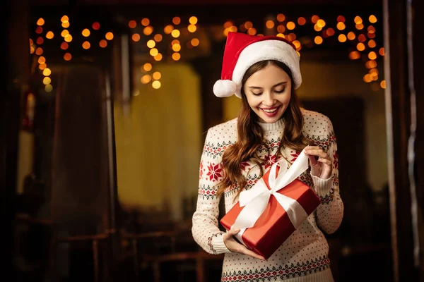 Здивована молода жінка в теплому светрі і капелюсі Санта відкриває подарункову коробку — стокове фото