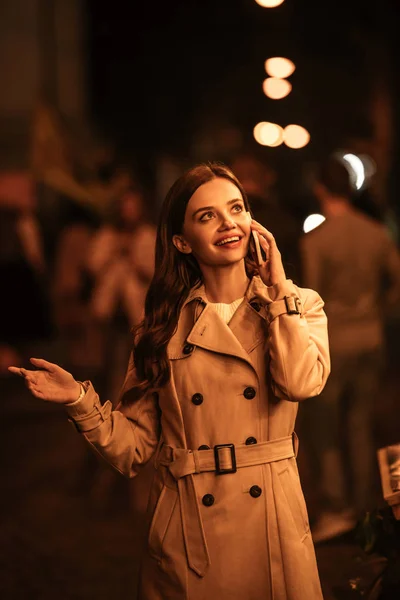 Hübsches, lächelndes Mädchen, das wegschaut, während es auf der Abendstraße mit dem Smartphone spricht — Stockfoto