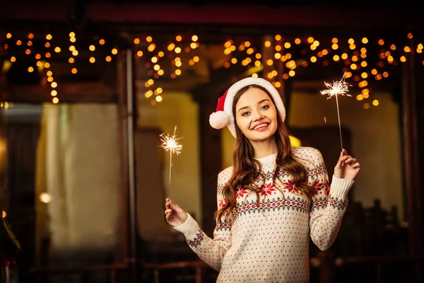 Fröhliche junge Frau in warmem Pullover und Weihnachtsmütze mit Wunderkerzen in der Hand, während sie in die Kamera lächelt — Stockfoto