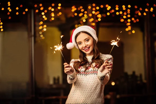 Happy young woman in warm sweater and santa hat holding sparklers while smiling at camera — Stock Photo