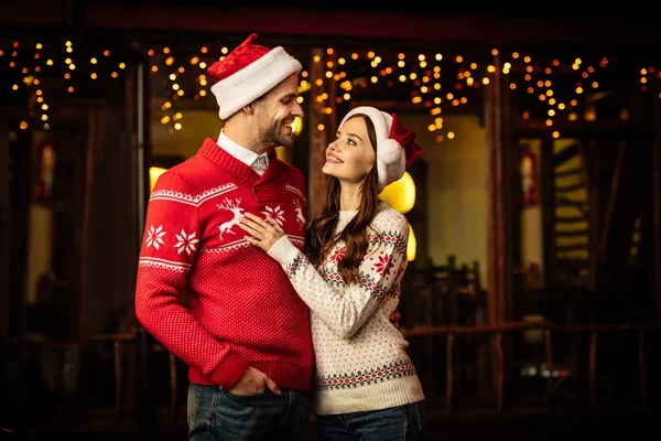 Glückliches junges Paar in warmen Pullovern und Weihnachtsmützen, das einander lächelnd ansieht — Stockfoto