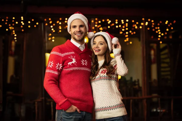 Cheerful young woman touching santa hat while embracing happy boyfriend — Stock Photo