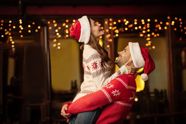 Cheerful young man holding happy girlfriend on hands while having fun on christmas eve — Stock Photo