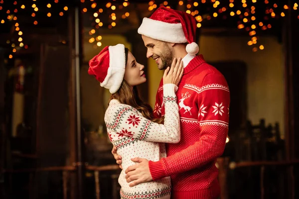 Feliz pareja joven en sombreros de santa y suéteres calientes abrazándose mientras se miran entre sí - foto de stock