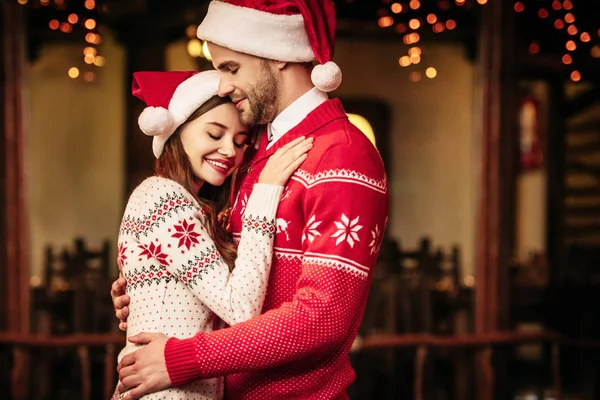 Glückliches junges Paar in Weihnachtsmützen und warmen Pullovern, das sich mit geschlossenen Augen umarmt — Stockfoto