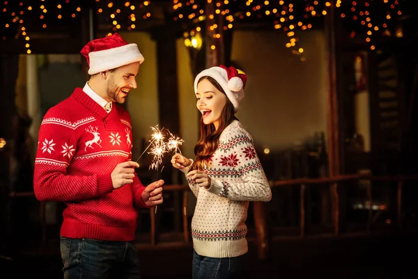 Fröhliches junges Paar in Weihnachtsmützen und warmen Pullovern mit Wunderkerzen an Heiligabend — Stockfoto