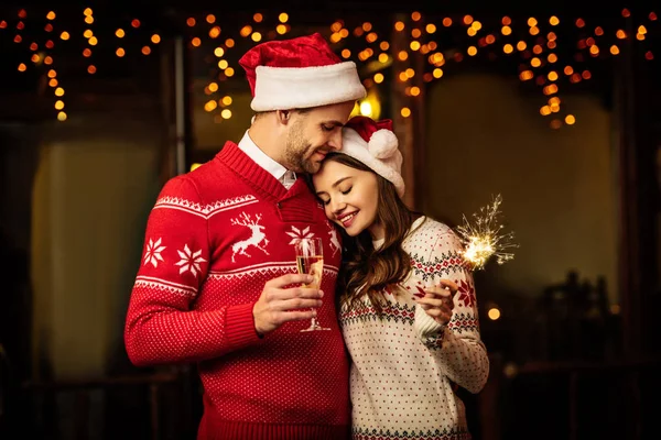 Homme souriant avec verre de champagne embrassant petite amie heureuse tenant scintillant — Photo de stock