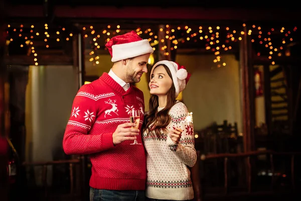 Glückliches junges Paar in Weihnachtsmannmützen, die einander beim Halten von Champagnerglas und Wunderkerze anschauen — Stockfoto