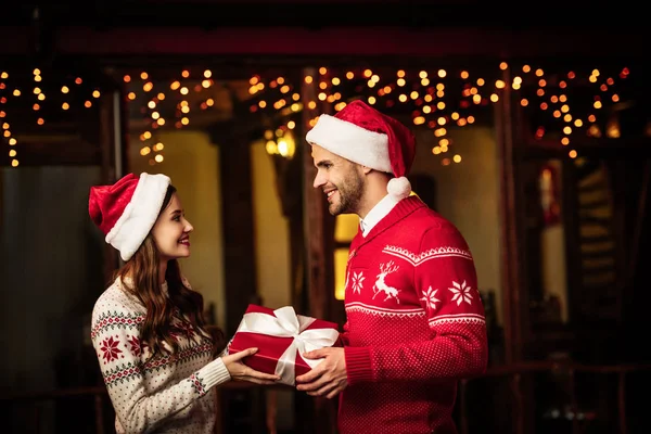 Glückliches junges Paar in warmen Pullovern und Weihnachtsmützen, die Geschenkschachtel in der Hand haltend und einander anschauend — Stockfoto