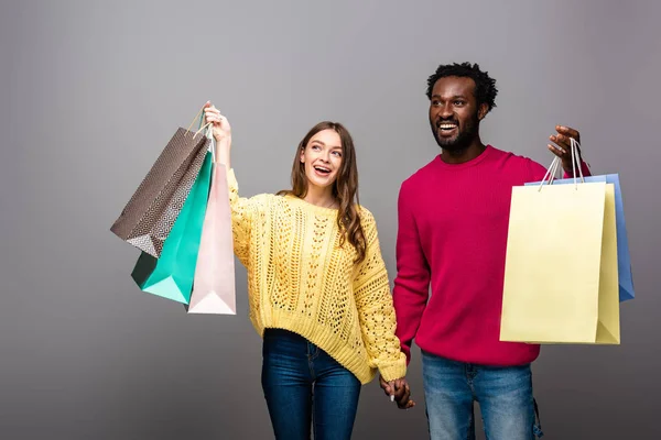 Feliz pareja interracial en suéteres tomados de la mano y bolsas de compras sobre fondo gris - foto de stock