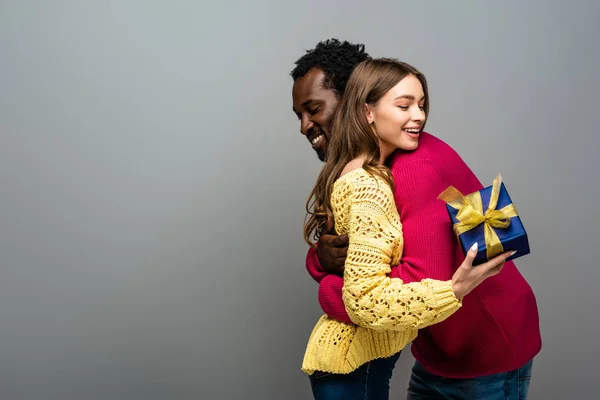 Feliz pareja interracial en suéteres sosteniendo presente y abrazando sobre fondo gris - foto de stock