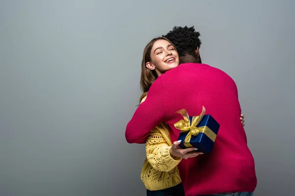 Feliz pareja interracial en suéteres sosteniendo presente y abrazando sobre fondo gris - foto de stock