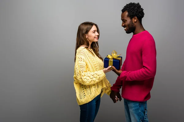 Feliz interracial pareja en suéteres celebración presente en gris fondo - foto de stock
