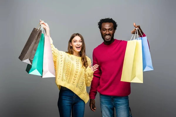 Feliz pareja interracial en suéteres tomados de la mano y bolsas de compras sobre fondo gris - foto de stock