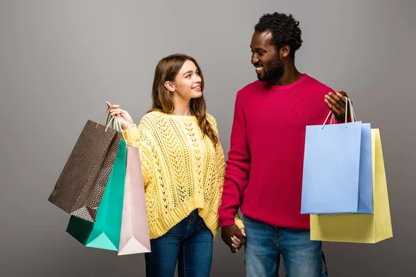 Feliz pareja interracial en suéteres tomados de la mano y bolsas de compras sobre fondo gris - foto de stock