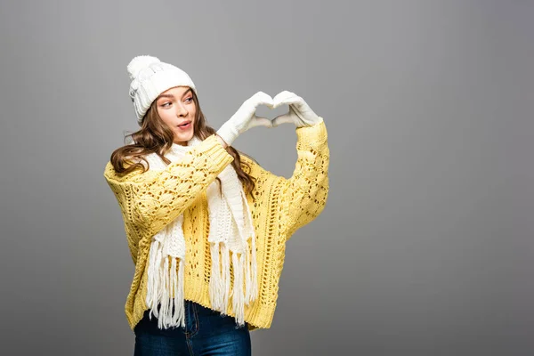 Happy girl in yellow sweater, scarf, hat and gloves showing heart gesture on grey — Stock Photo