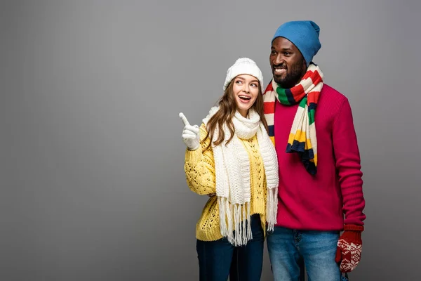 Sonriente pareja interracial en invierno traje abrazando y señalando con el dedo sobre fondo gris - foto de stock