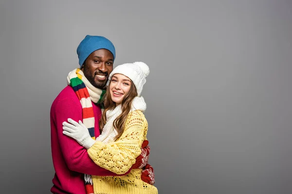 Усміхнена міжрасова пара в зимовому вбранні обіймається на сірому фоні — стокове фото