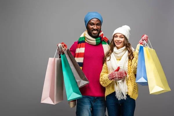 Glückliches gemischtrassiges Paar im winterlichen Outfit mit Einkaufstaschen auf grauem Hintergrund — Stockfoto