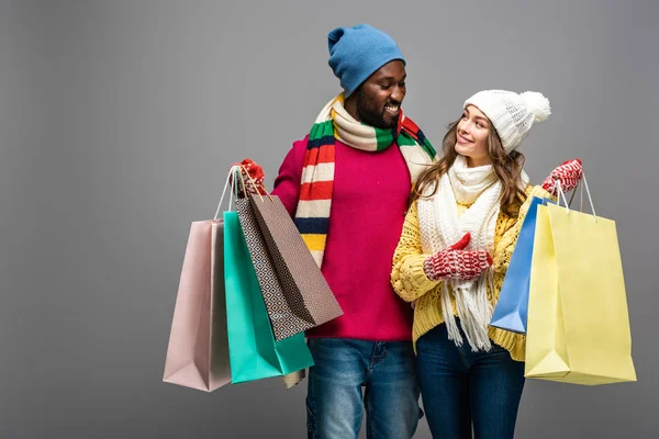 Happy interracial couple in winter outfit holding shopping bags on grey background — Stock Photo