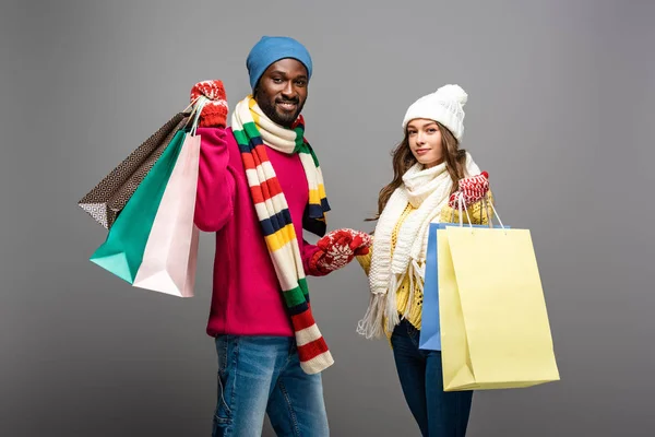 Glückliches gemischtrassiges Paar im winterlichen Outfit mit Einkaufstaschen auf grauem Hintergrund — Stockfoto