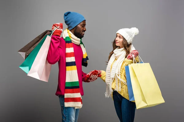 Sonriente pareja interracial en invierno traje sosteniendo bolsas de compras sobre fondo gris - foto de stock