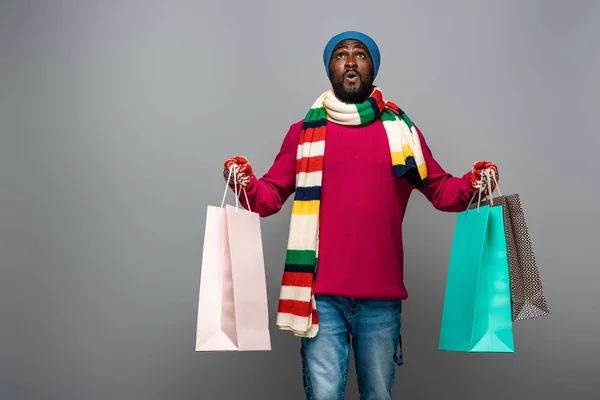 Shocked african american man in winter outfit with shopping bags on grey background — Stock Photo
