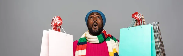 Sorprendido hombre afroamericano en traje de invierno con bolsas de compras aisladas en gris, tiro panorámico - foto de stock