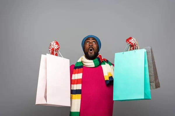 Shocked african american man in winter outfit with shopping bags on grey background — Stock Photo