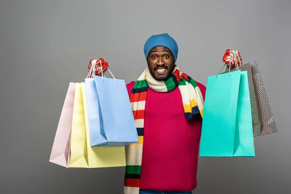 Hombre afroamericano feliz en traje de invierno con bolsas de compras sobre fondo gris - foto de stock