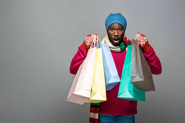 Sorprendido hombre afroamericano en traje de invierno con bolsas de compras sobre fondo gris - foto de stock